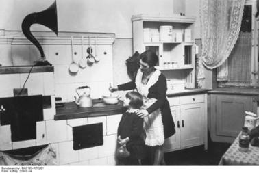 Frau mit Kind beim Kochen und über ein Grammophon Radio hörend, 1925
